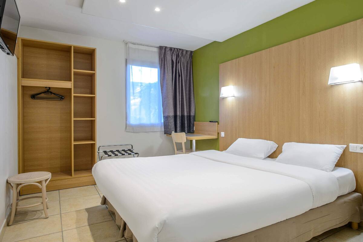 A hotel room with a large bed, a wooden wardrobe, a small table, and a chair.