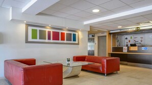 A reception area with a glass-top table, red leather sofas, and a wall-mounted art piece. - B&B HOTEL Nîmes Ville Active (Nîmes)