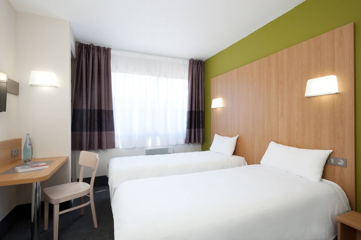 A hotel room with two beds, a desk with a chair, a window with curtains, and wall-mounted lights.