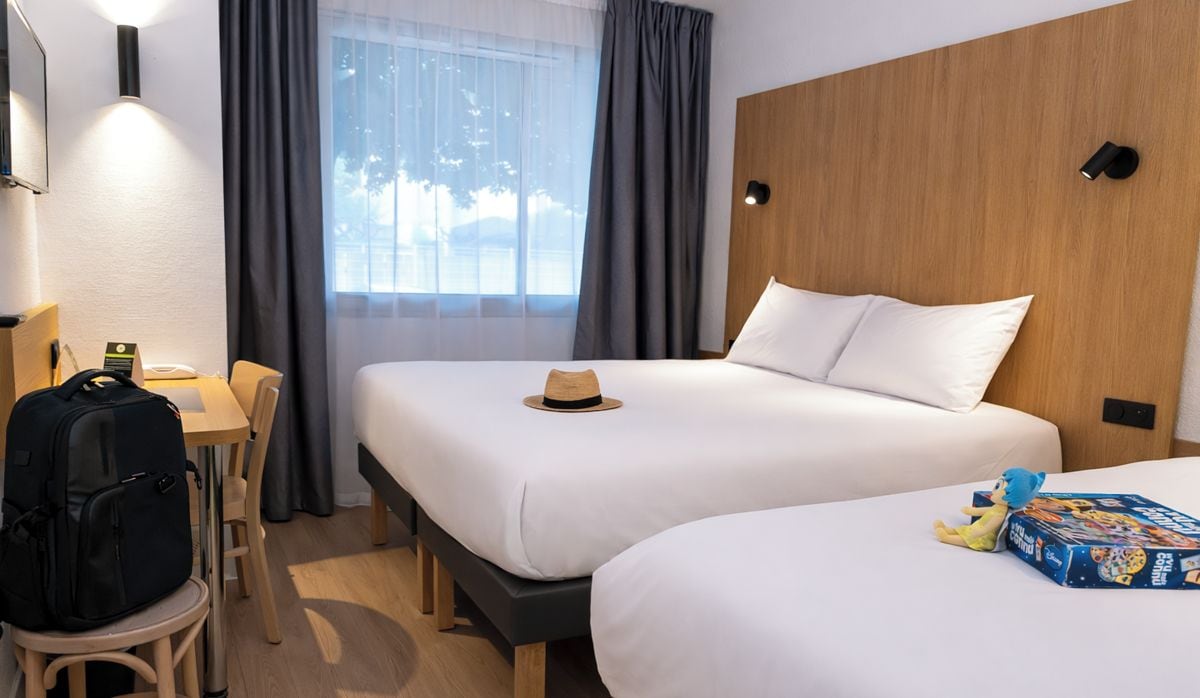 Une chambre d’hôtel avec deux lits, un bureau, une chaise, une valise et une fenêtre avec des rideaux.