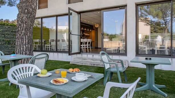 An outdoor dining area with tables and chairs, a tree, and a building entrance. - B&B HOTEL Nîmes Ville Active (Nîmes)