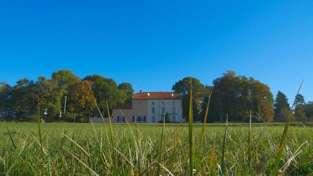 Une grande maison avec un toit rouge, entourée d’un champ verdoyant et d’arbres.