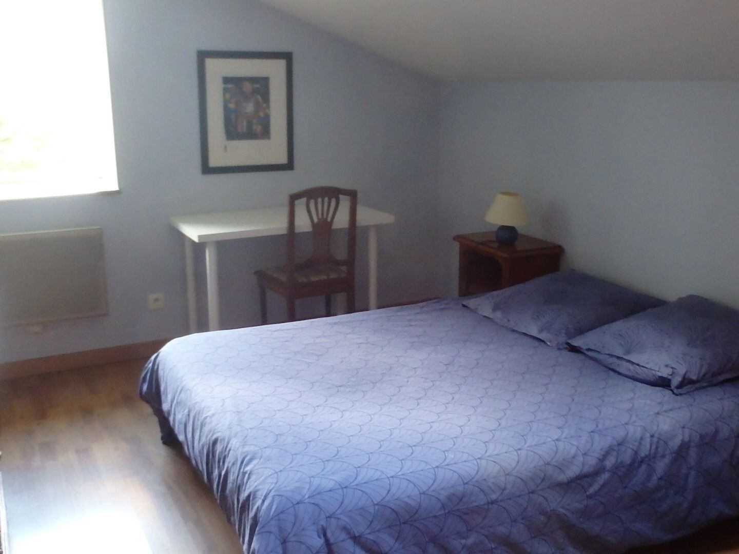 A bedroom with a bed, a desk, a chair, and a nightstand with a lamp.