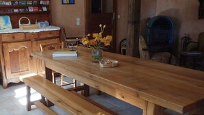 Una mesa de madera para comer con bancos, un jarrón con flores y una estantería en una habitación con suelo de piedra.
