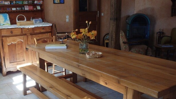 Une table à manger en bois avec des bancs, un vase rempli de fleurs et une bibliothèque dans une pièce dont le sol est recouvert de pierres.