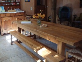 A wooden dining table with benches, a vase with flowers, and a bookshelf in a room with stone flooring.