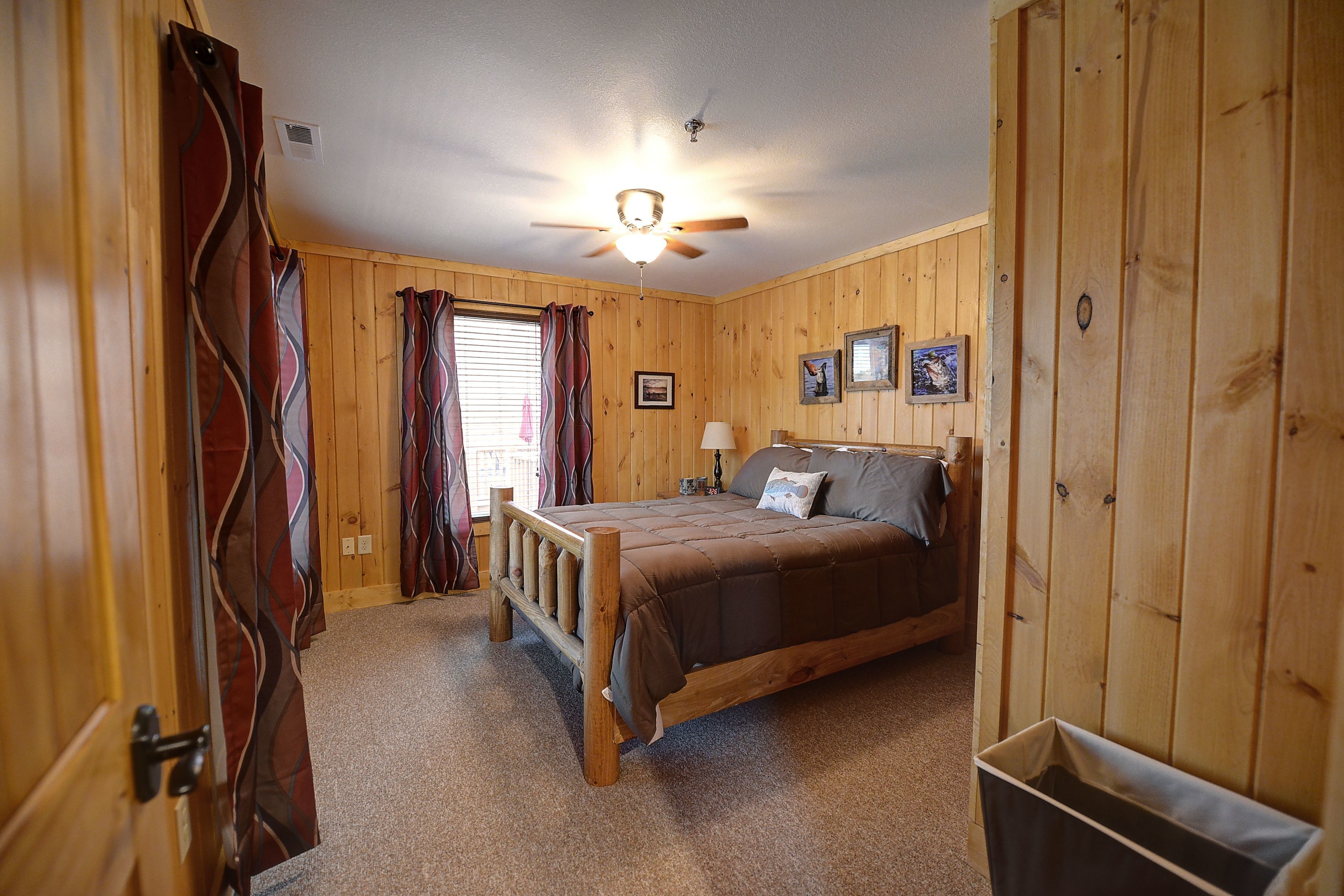 Een slaapkamer met houten lambrisering, een bed, gordijnen, een plafondventilator en een prullenbak.