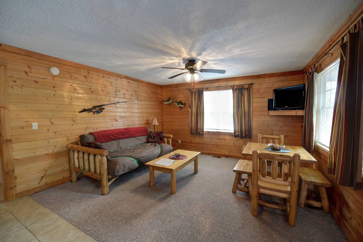 Ein Urigen Holzblockhaus-Interieur mit Deckenventilator, einer Couch, einem Couchtisch, einem Esstisch mit Stühlen und einem an der Wand montierten Fernseher.