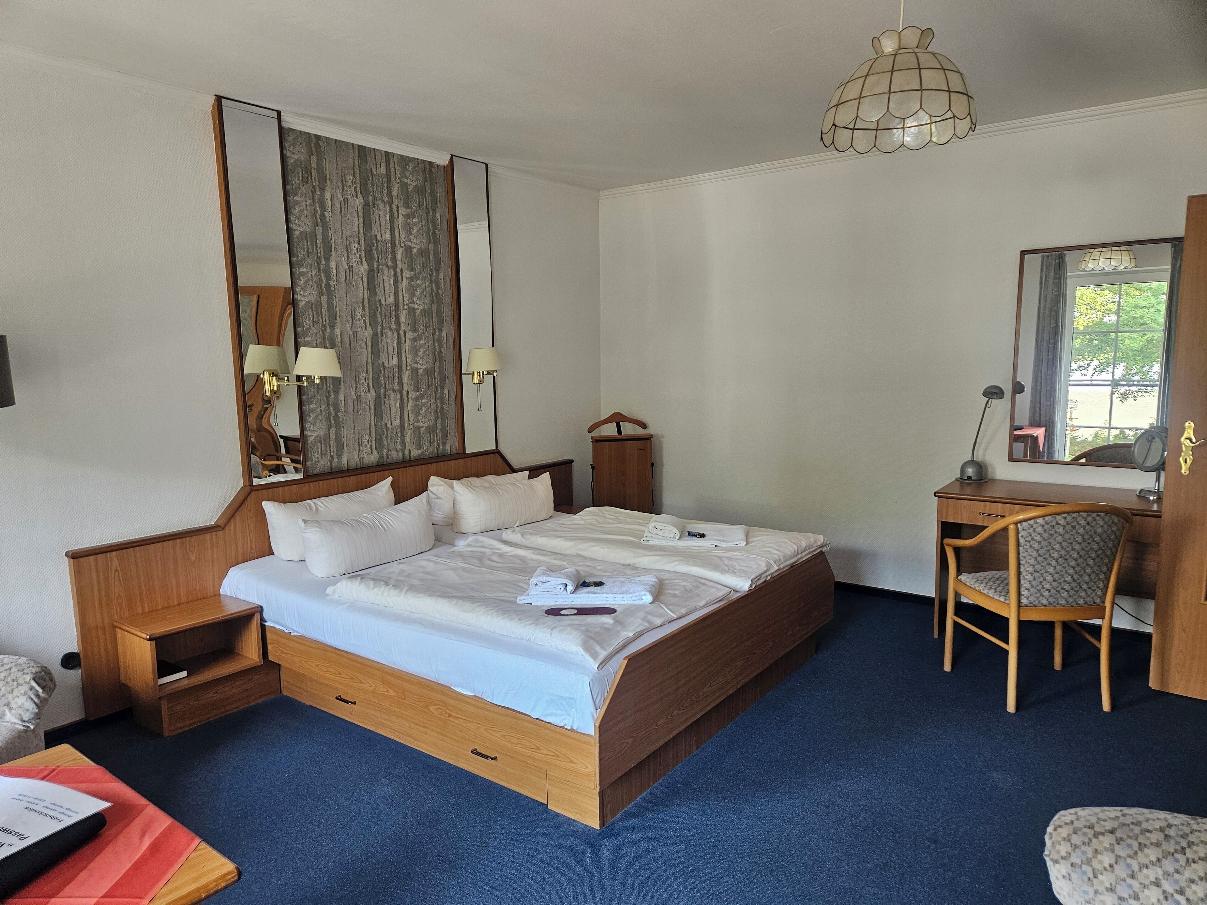 Habitación de hotel con una cama grande, un escritorio de madera, una silla, un espejo y una lámpara colgante.