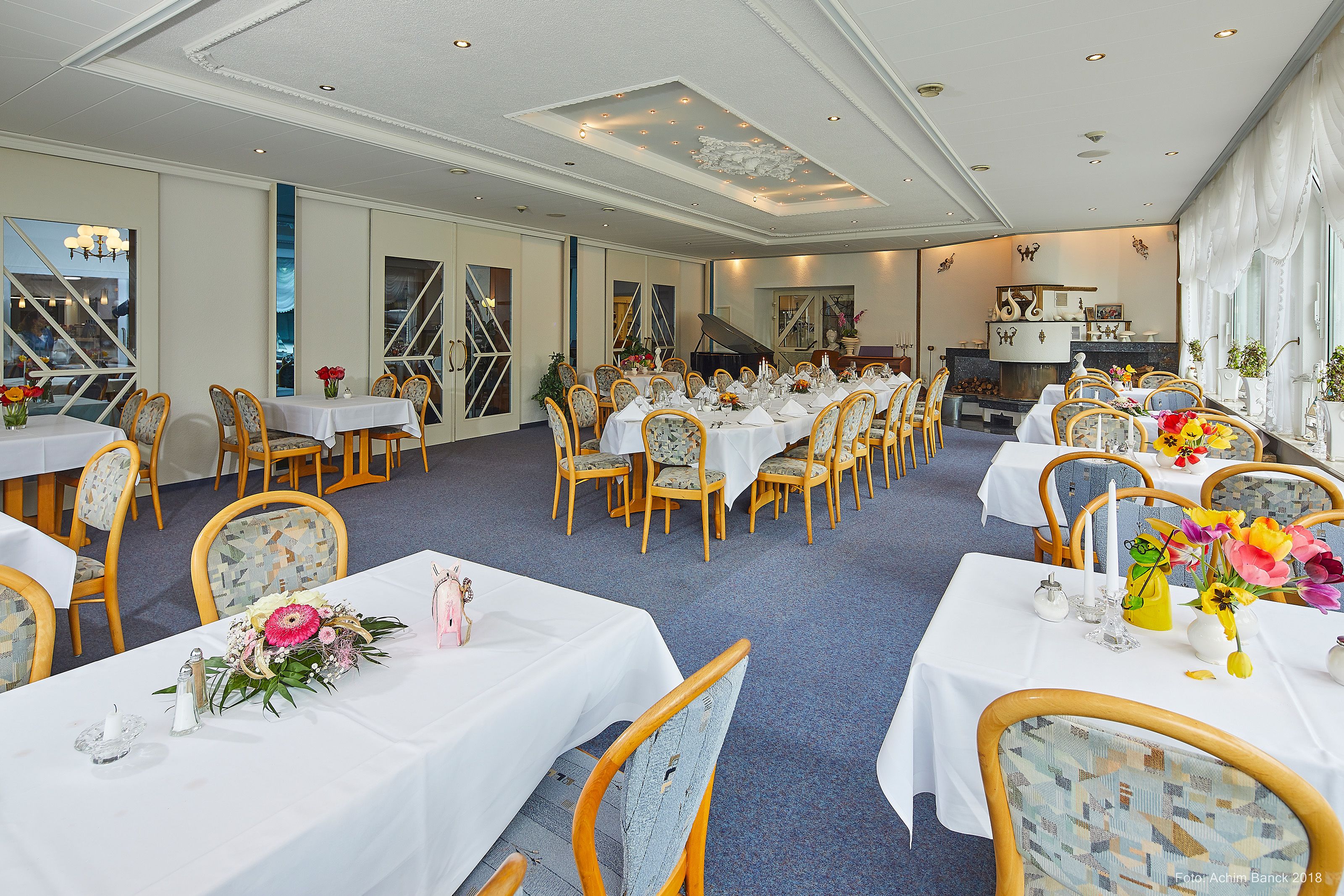 A spacious dining area with multiple tables and chairs, large windows, and a piano in the background.
