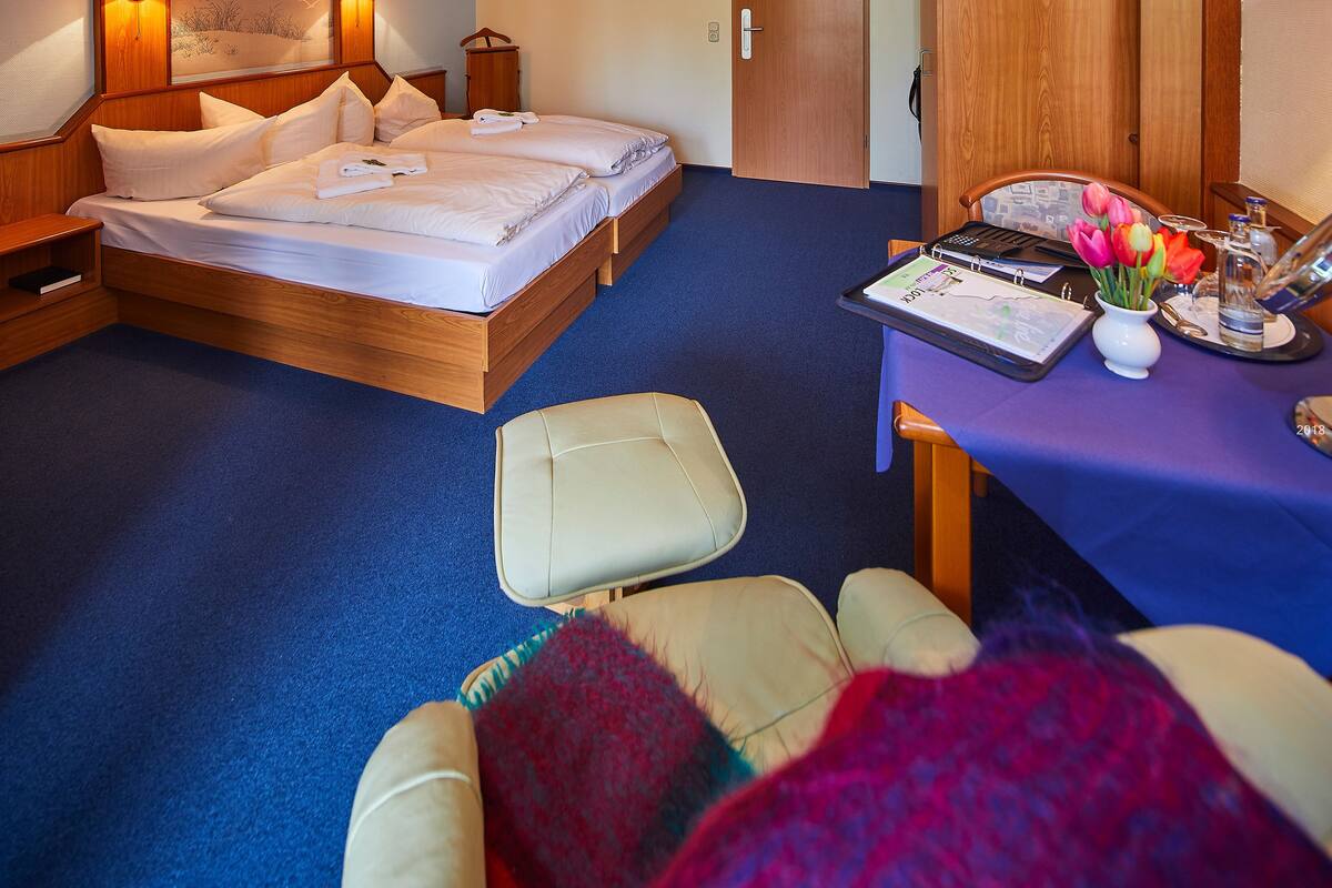 A hotel room with a double bed, a desk with a laptop, a chair, a small table with a vase of flowers, and a wooden wardrobe.
