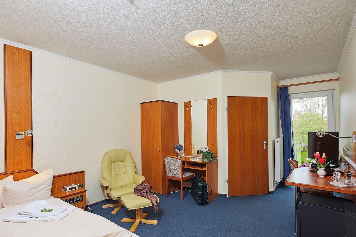 A hotel room with a bed, a desk with a chair, a wardrobe, a mirror, and a window with curtains.