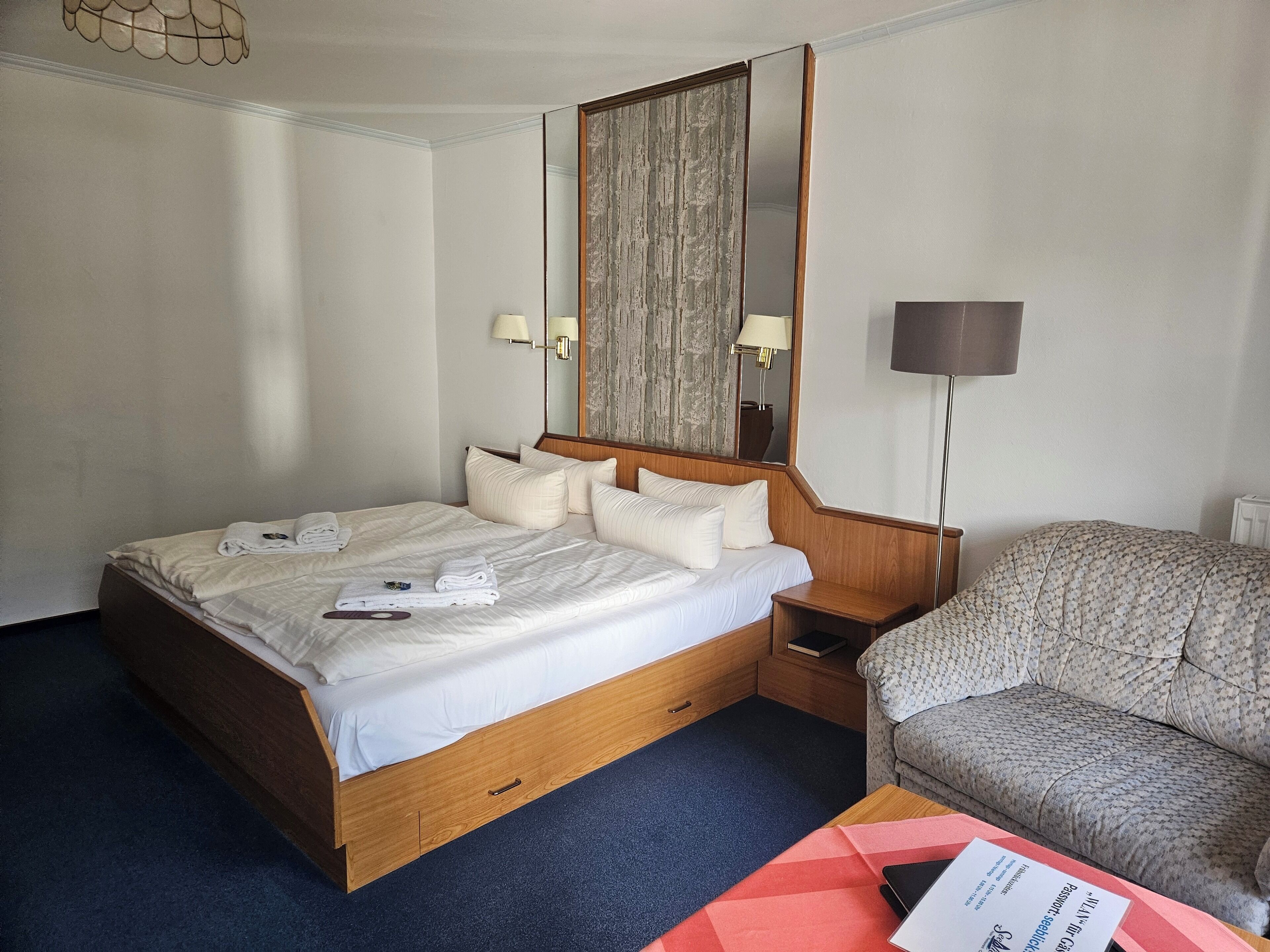 Una habitación de hotel con cama, cabecera de madera, un espejo grande, mesita de noche, lámpara, sofá y piso alfombrado.