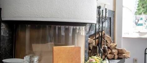 Una chimenea moderna con detalles decorativos blancos y dorados, una placa de madera y un florero con flores sobre una encimera de mármol.