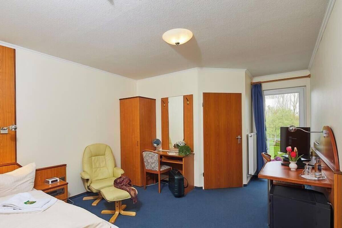 A hotel room with a bed, a desk with a chair, a wardrobe, a mirror, and a window with curtains.