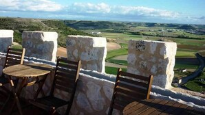 Una terraza con muros de piedra y sillas de madera, con vistas a un paisaje rural con campos y un camino sinuoso.