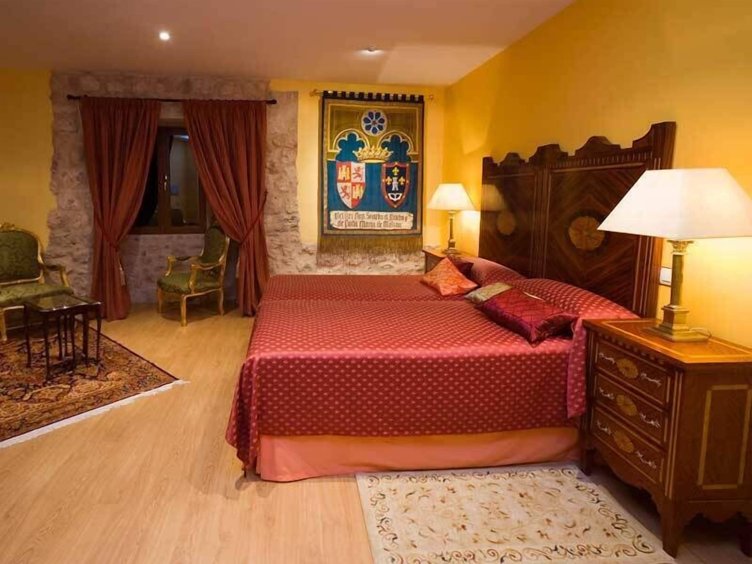 A bedroom with a large bed, two chairs, a wooden dresser, a patterned rug, and a wall with a coat of arms.
