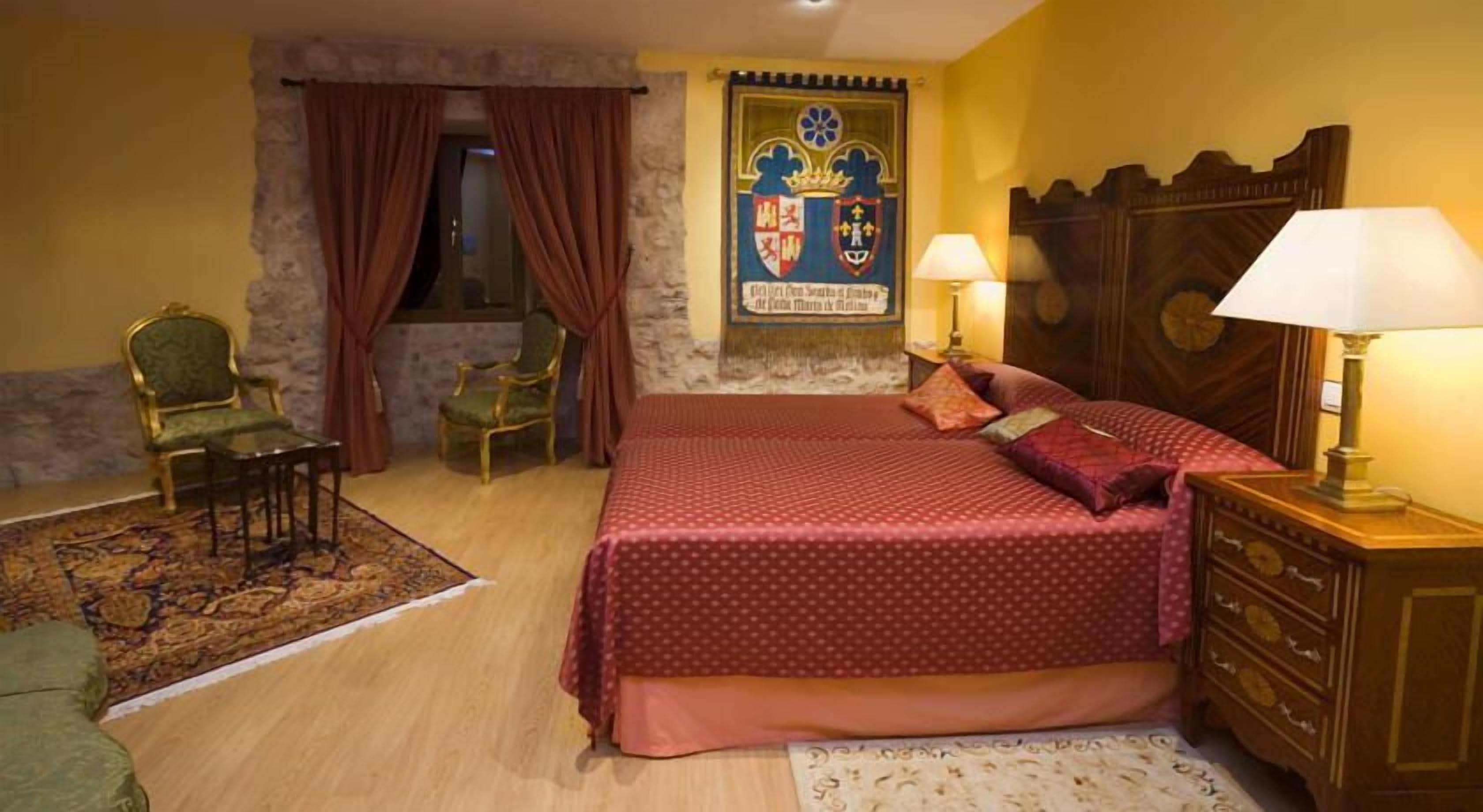 Un dormitorio con cama, dos sillas, un aparador de madera, una mesita de noche con lámpara, una ventana con cortinas y un escudo en la pared.