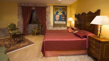 Un dormitorio con cama, dos sillas, un aparador de madera, una mesita de noche con lámpara, una ventana con cortinas y un escudo en la pared.