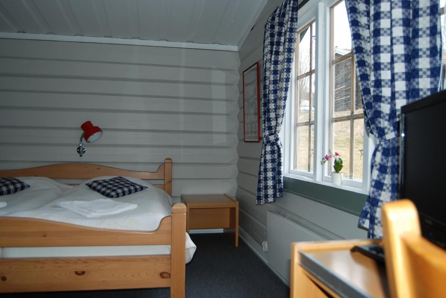 Un dormitorio con una cama de madera, una mesita de noche pequeña, una lámpara roja y una ventana con cortinas a cuadros.