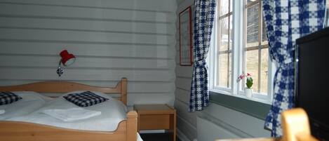 Un dormitorio con una cama de madera, una mesita de noche pequeña, una lámpara roja y una ventana con cortinas a cuadros.