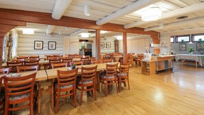 A spacious dining area with wooden tables and chairs, a kitchen area, and a view of the outdoors. - Dala Wärdshus (Rattvik)