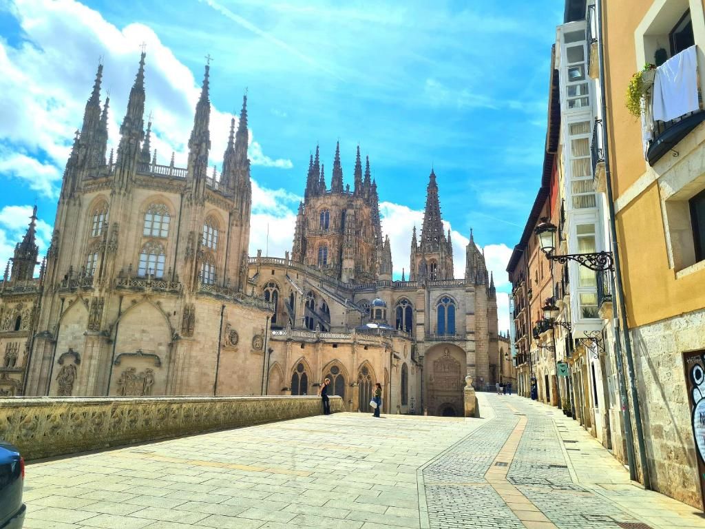 Una catedral amb una gran agulla central i diverses agulletes més petites, una plaça pavimentada al primer terme i edificis amb balcons a la dreta.
