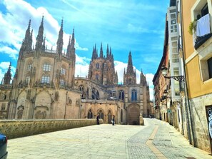 Una catedral con una alta aguja central y varias agujas más pequeñas, una plaza adoquinada al frente y edificios con balcones a la derecha.
