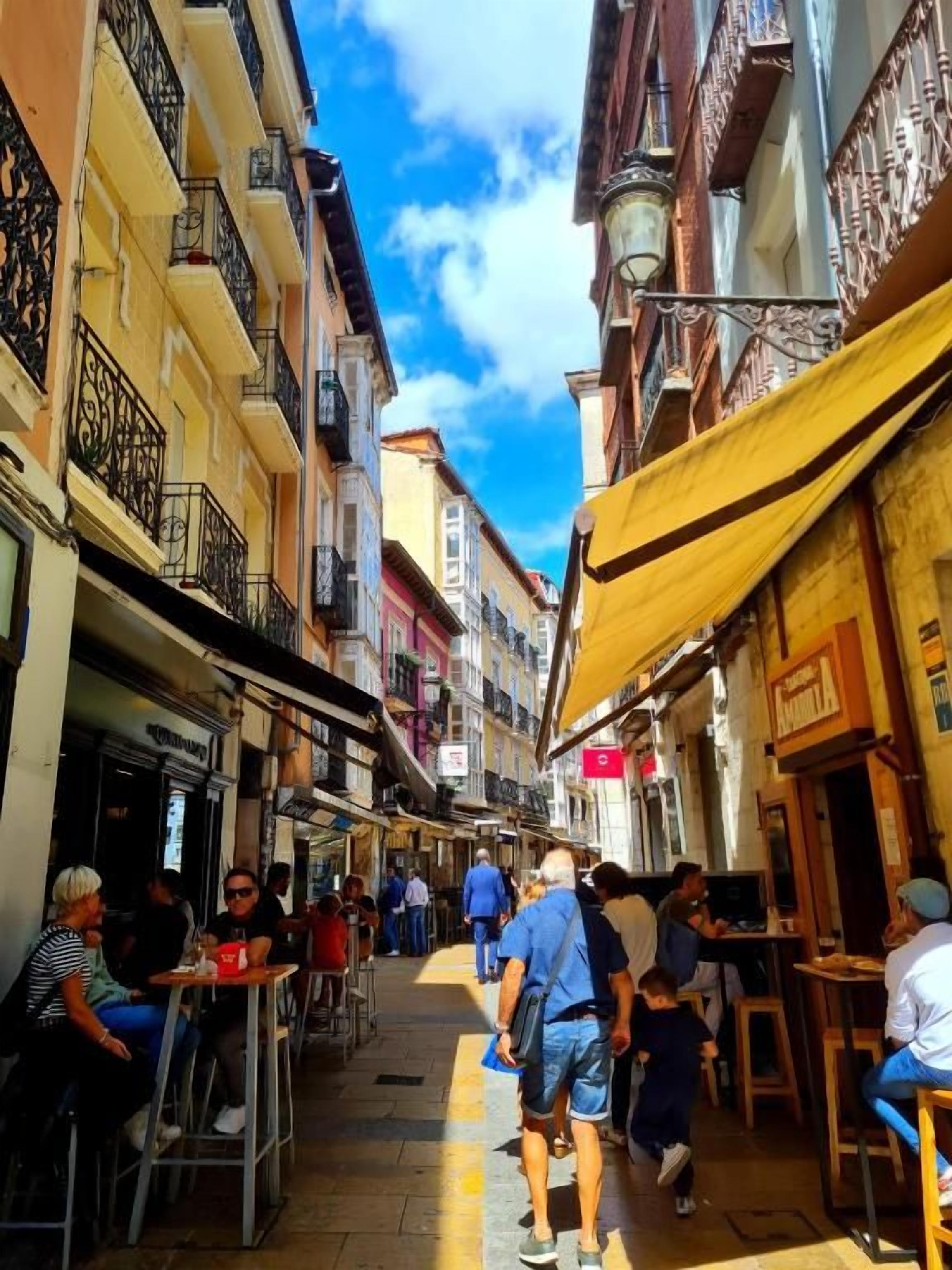 Una calle estrecha con terrazas al aire libre, edificios con balcones y cielo despejado.