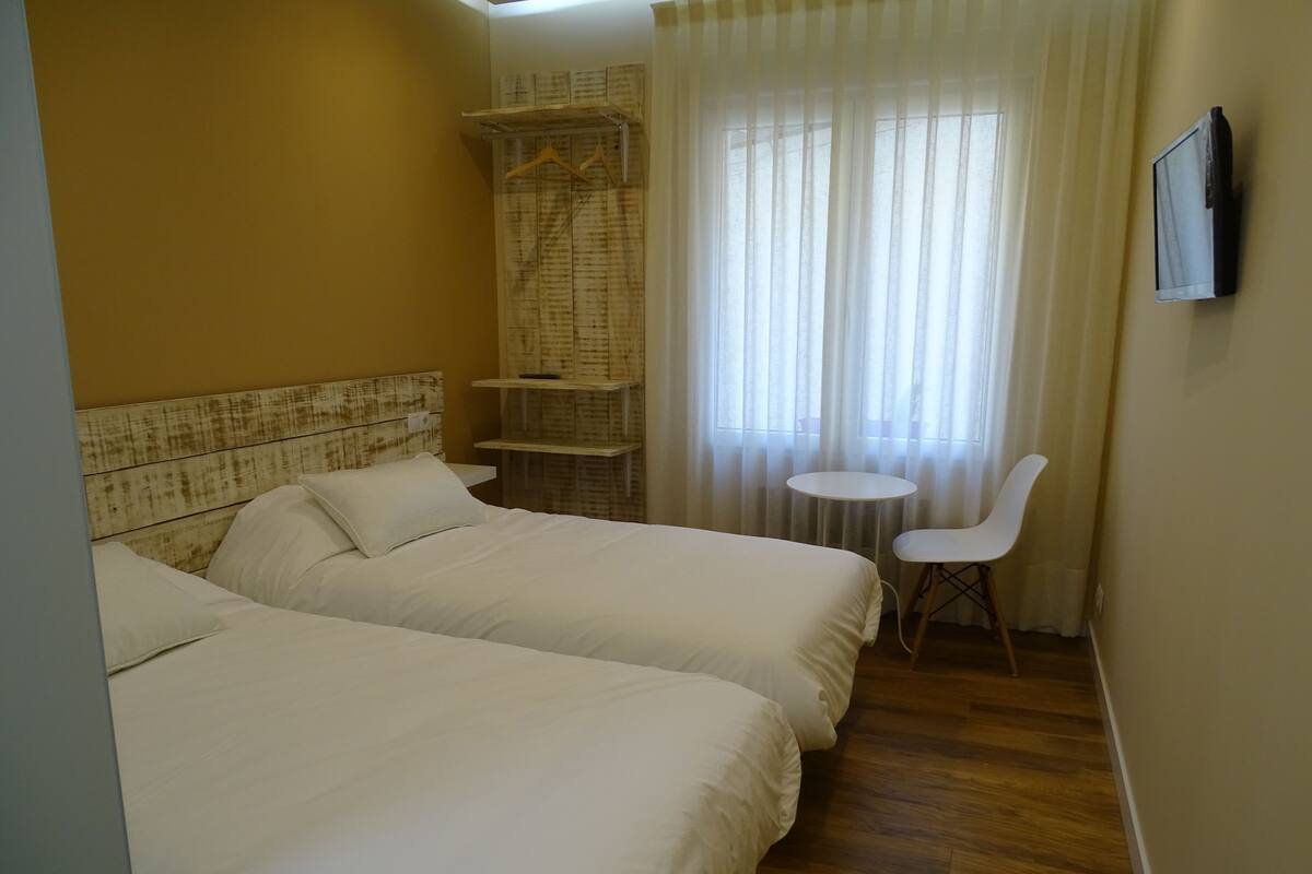 A hotel room with two beds, a small table, a chair, and a window with curtains.