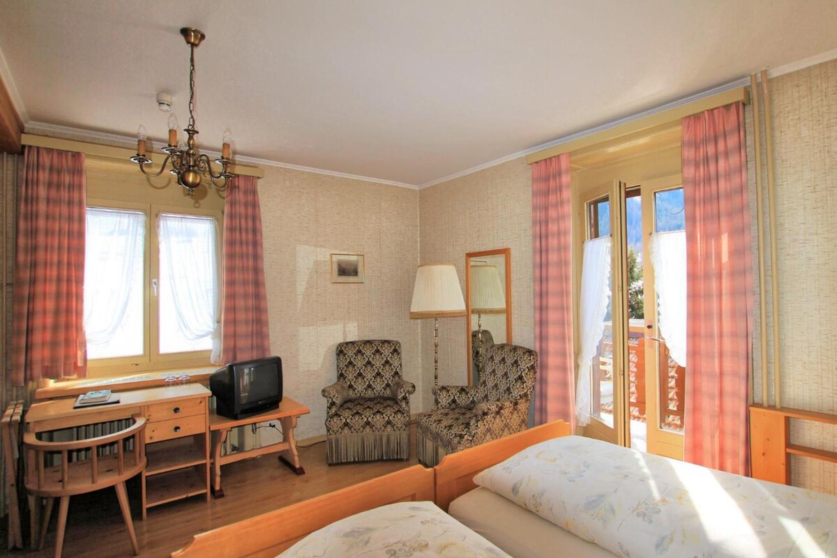 Habitación de hotel con una cama, un escritorio con televisor, dos sillones, una lámpara y una ventana con cortinas.