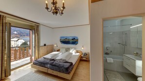 A bedroom with a wooden bed, a view of the outdoors, a chandelier, and a bathroom with a bathtub and mirror. - Hotel Restaurant Schweizerhof (Val Muestair)