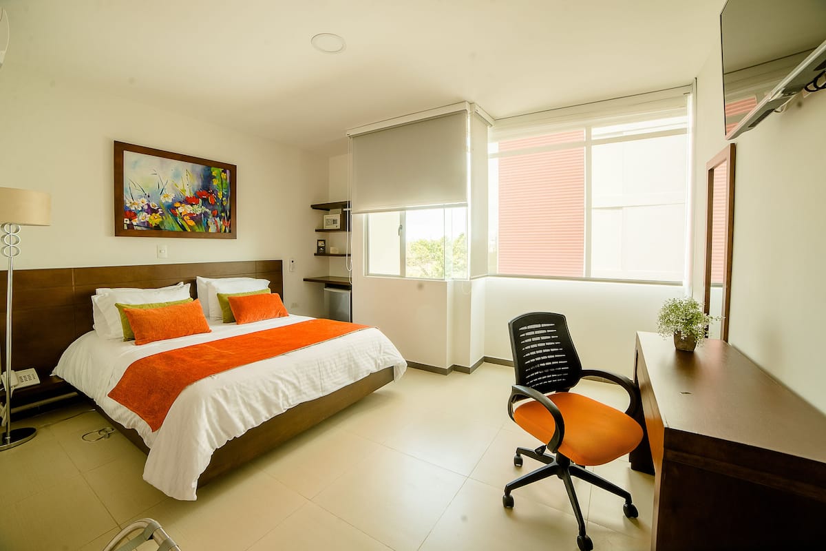 Una habitación de hotel con cama, escritorio, silla, ventana y un cuadro en la pared.