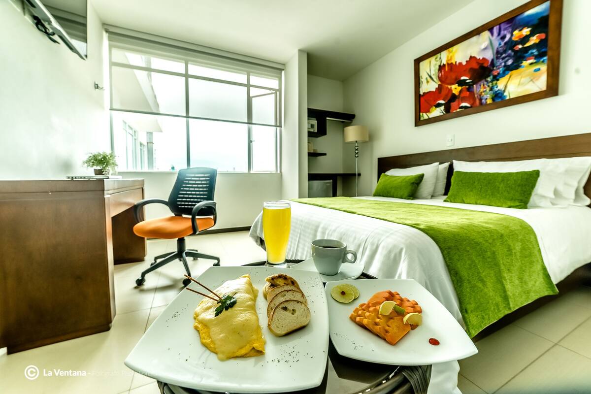 Habitación de hotel con cama, escritorio, silla, mesa de desayuno y ventana con vistas.