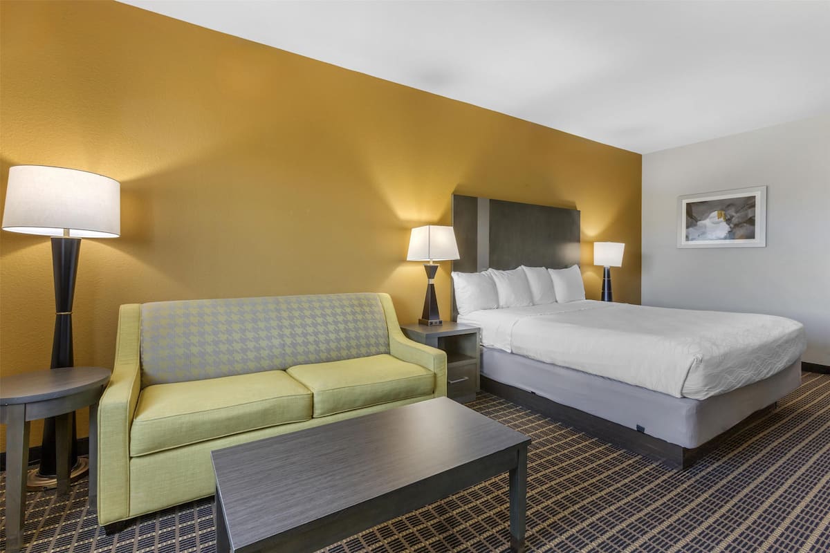 Habitación de hotel con pared de acento amarilla, cama, sofá, dos mesitas de noche con lámparas y un cuadro en la pared.