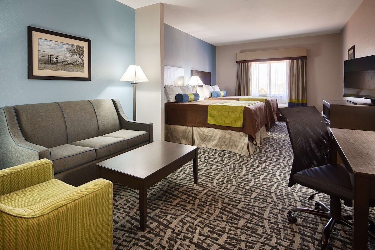 A hotel room with a sofa, armchair, desk, and a bed with multiple pillows.