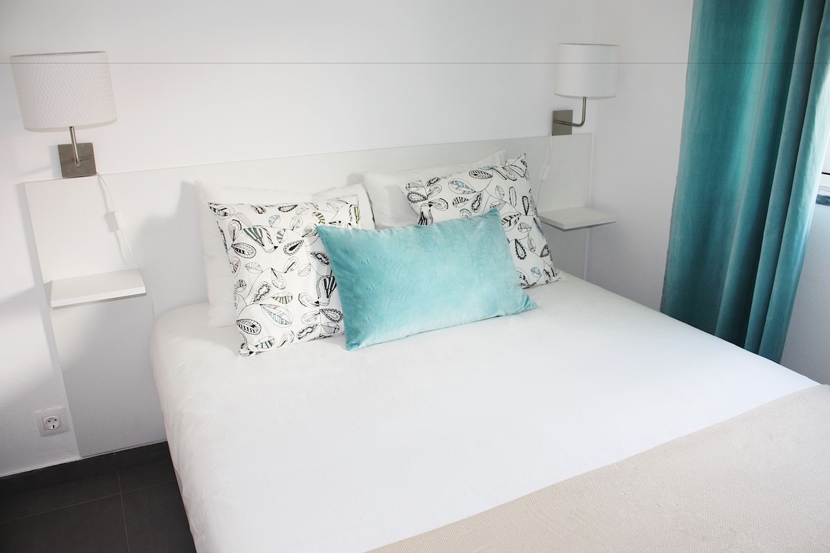 Habitación de hotel con una cama bien hecha, almohadas estampadas, un cojín de color verde azulado y dos lámparas fijadas a la pared.
