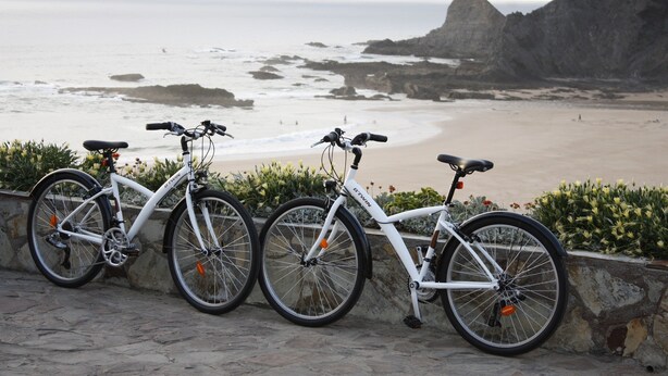 Una bicicleta blanca estacionada junto a un muro de piedra con vista a una playa y acantilados rocosos.