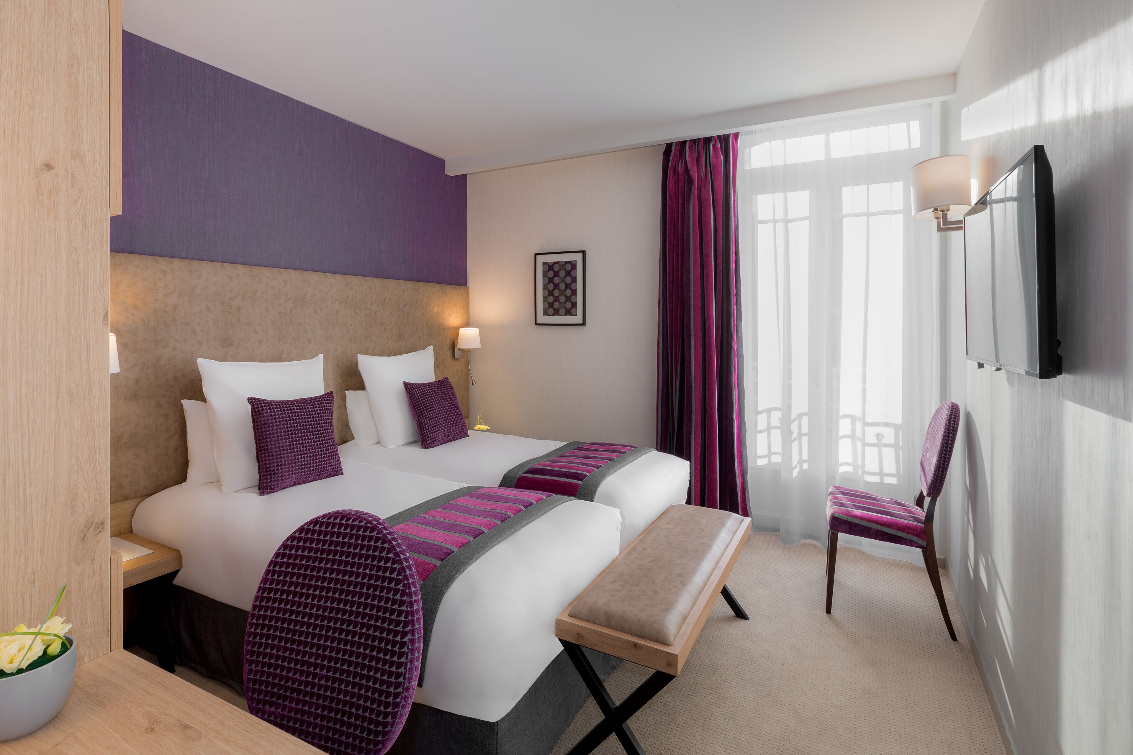 Ein Hotelzimmer mit zwei Betten, einer Bank, einem Sessel, einem Fernseher und einem Fenster mit Vorhängen.