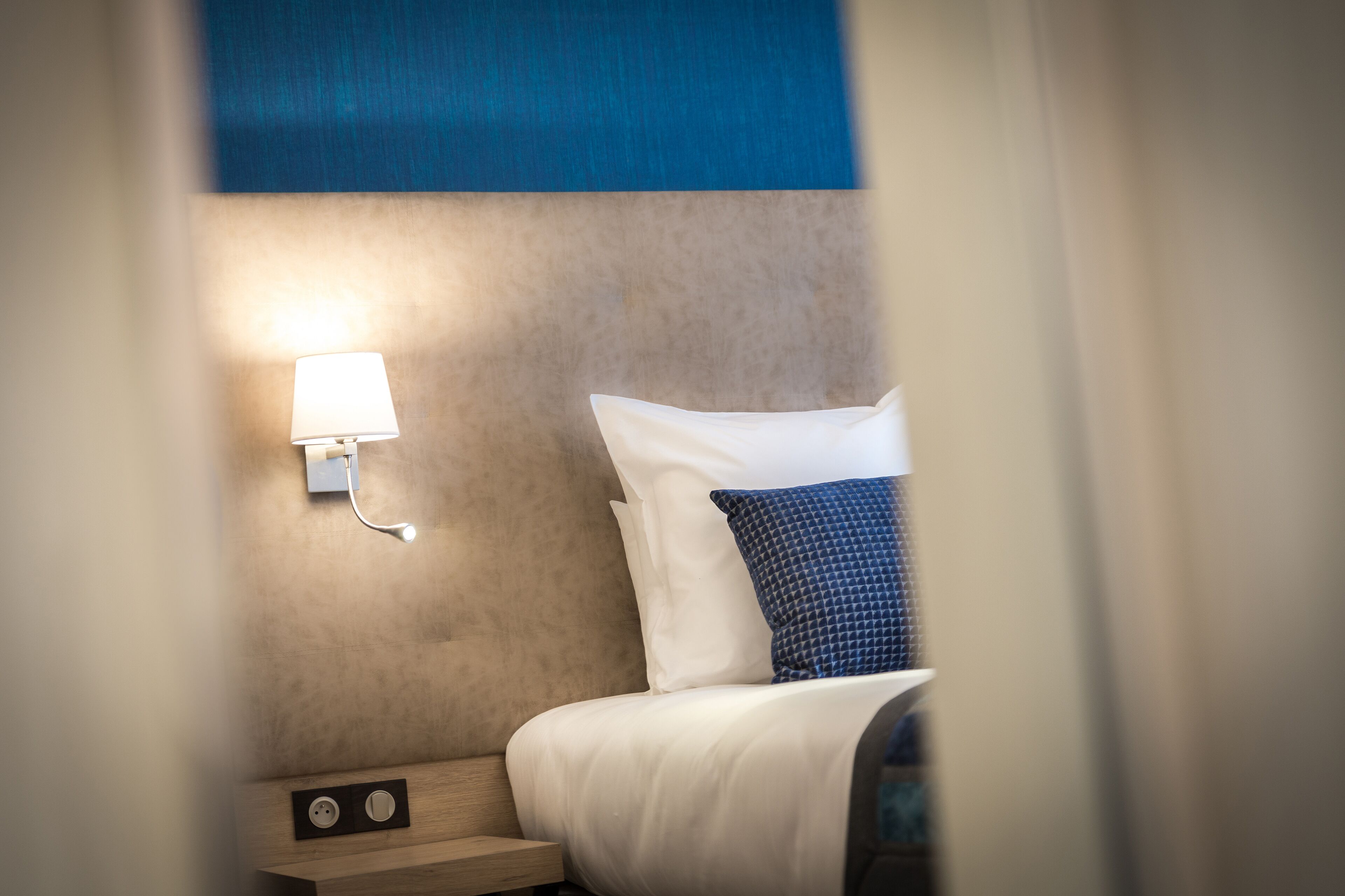 Habitación de hotel con cama, mesita de noche, lámpara de pared y toma de corriente.