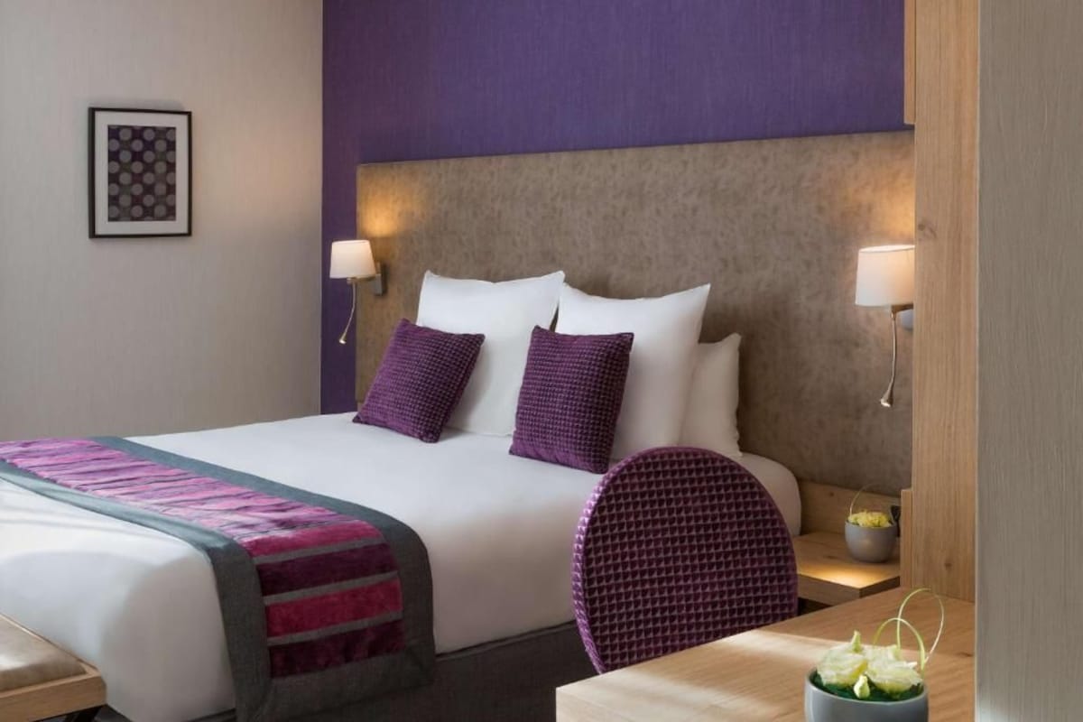 Ein Hotelzimmer mit einem Bett, zwei Kissen, einem Nachttisch, einem Stuhl und einem kleinen Tisch mit einer Pflanze.