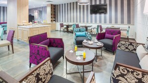 Un hall d’hôtel moderne doté d’un bar central, de différents espaces aménagés pour s’asseoir et d’un téléviseur fixé au mur.