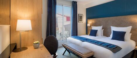 A hotel room with a bed, bedside tables, a desk, a chair, a lamp, and a window with curtains.