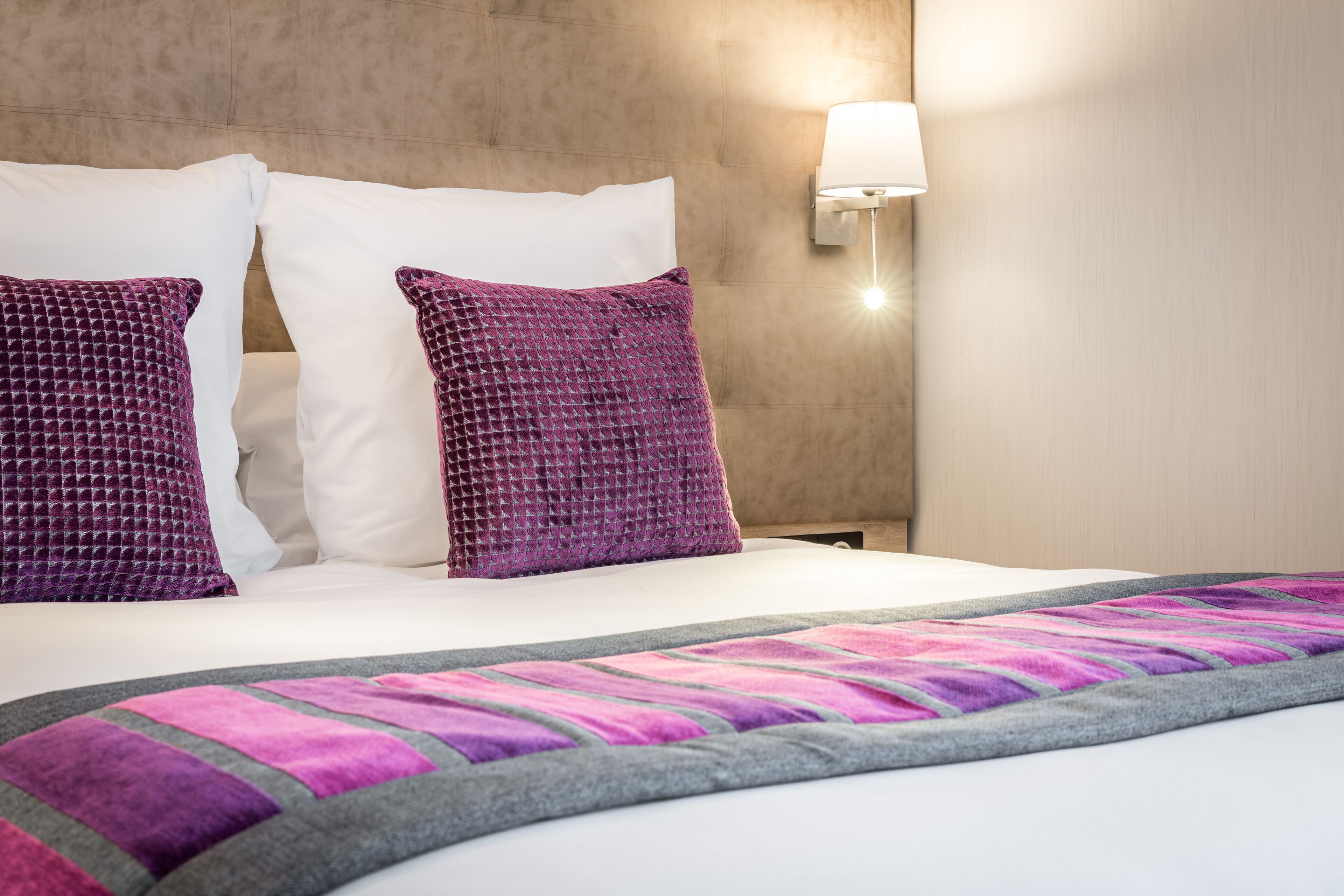 A neatly made bed with white and purple pillows, a striped blanket, and a wall-mounted lamp.
