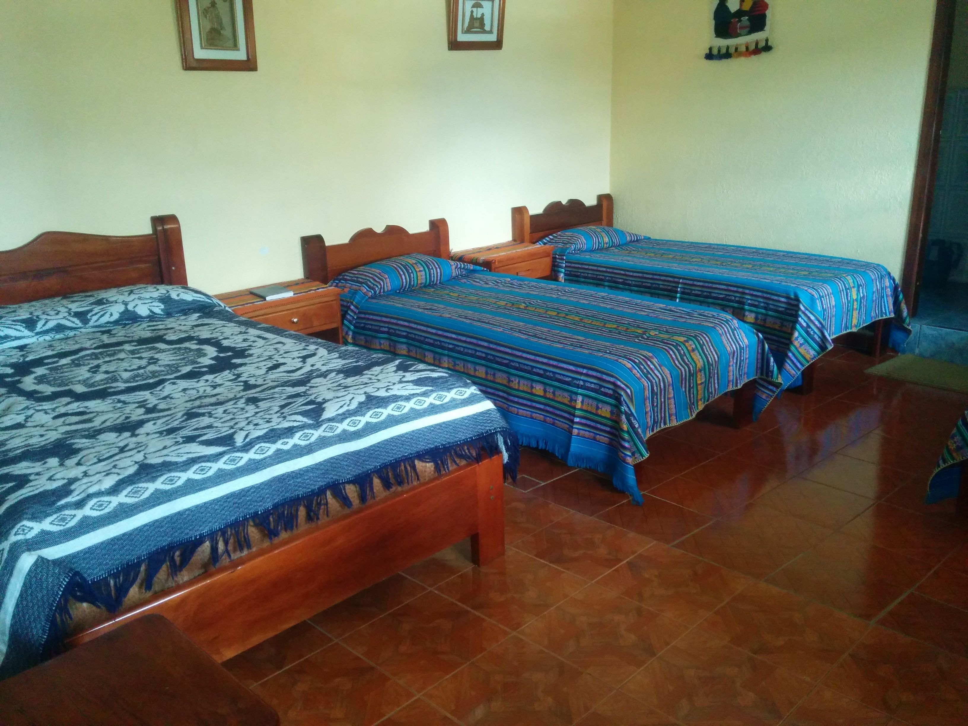 A room with three wooden beds, each with a patterned bedspread. The room has tiled flooring and plain walls with a few framed pictures.