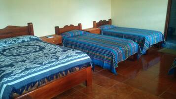 A room with three wooden beds, each with a patterned bedspread. The room has tiled flooring and plain walls with a few framed pictures.