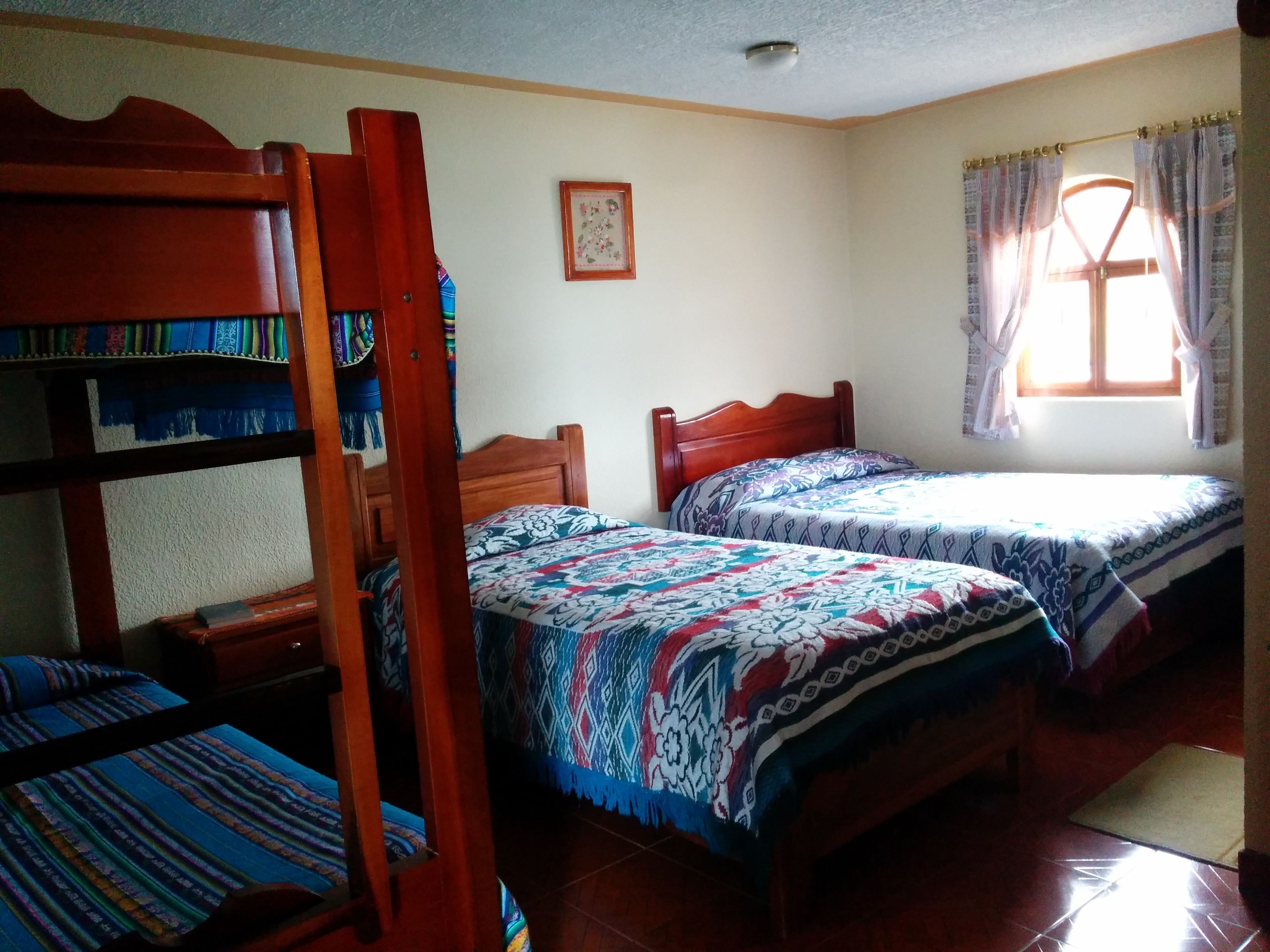 A dormitory room with bunk beds and a single bed, a window with curtains, and a framed picture on the wall.