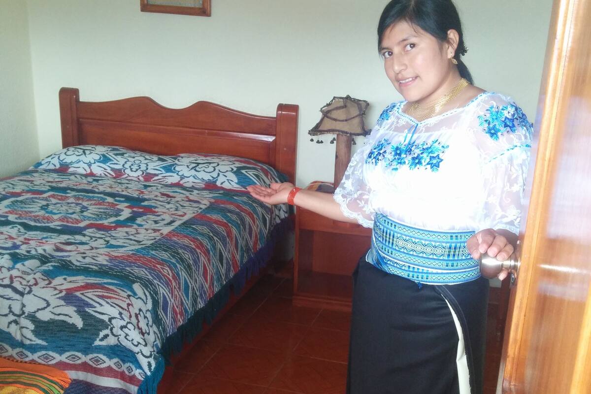 A room with a wooden bed, a patterned bedspread, a small table with a lamp, and a wall-mounted picture.