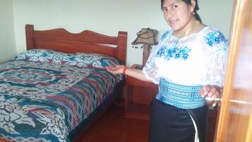 A room with a wooden bed, a patterned bedspread, a small table with a lamp, and a wall-mounted picture.