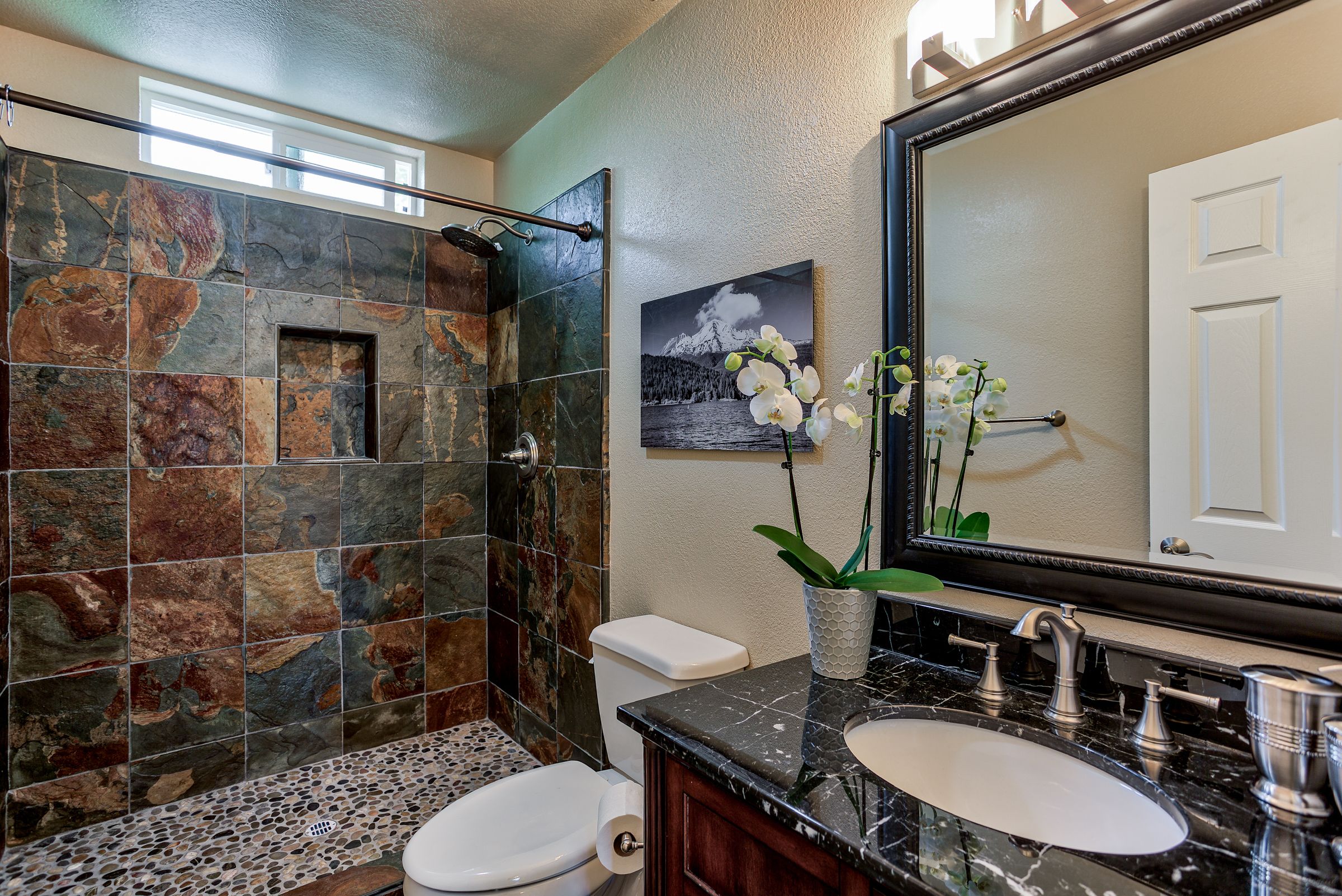 A bathroom with a stone shower, a toilet, a sink with a marble countertop, and a mirror above the sink.